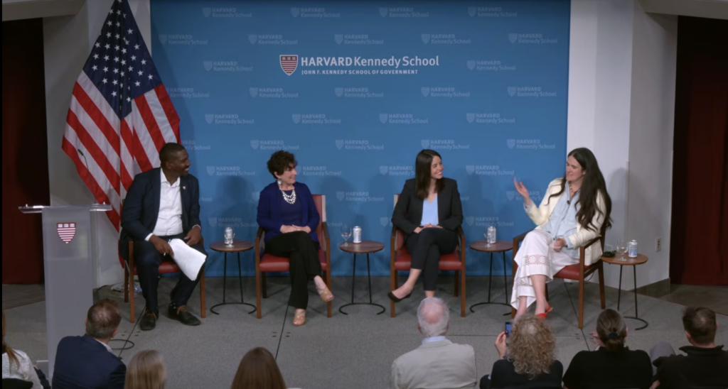 Four people, including Micaela, our CEO and Co-Founder, sit on a panel. The panelists are of different races and genders and are wearing business attire. They’re sitting in wooden arm chairs with the American flag hanging on a pole behind them next to a blue backdrop with the Harvard Kennedy School logo patterned on it. Three panelists are turned towards Micaela as she speaks.